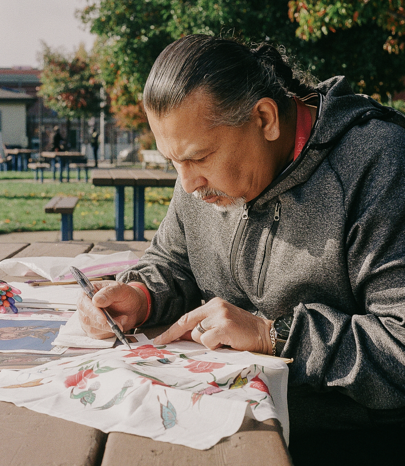 An older man with long hair in a grey hoodie sitting on a park bench painting. 